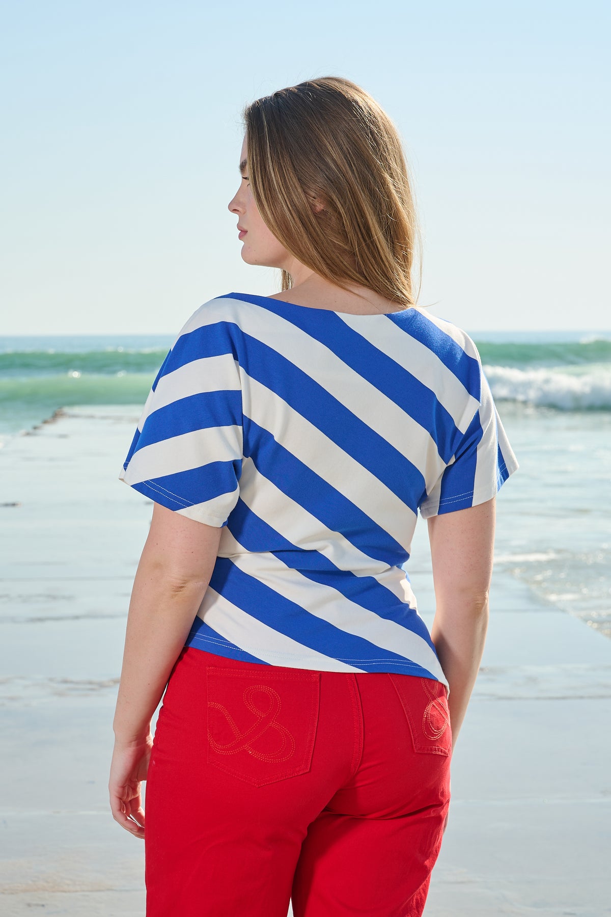 A woman standing on a beach, shown from the back, wearing a blue and white diagonally striped short-sleeve top and bright red pants.