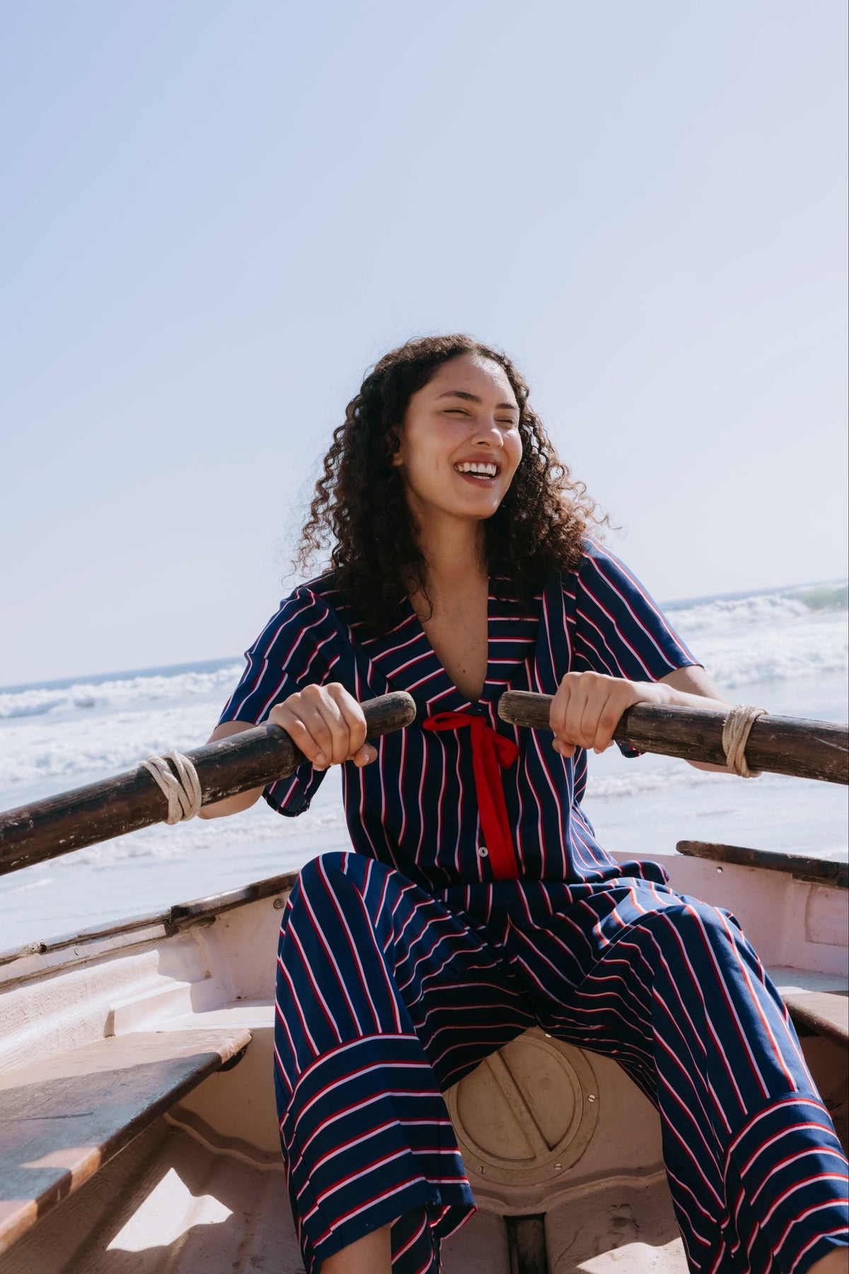A woman with curly dark hair sits in a rowboat on the water, wearing a matching navy blue striped set with red and navy pinstripes and a red tie at the chest