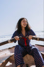 A woman with curly dark hair sits in a rowboat on the water, wearing a matching navy blue striped set with red and navy pinstripes and a red tie at the chest