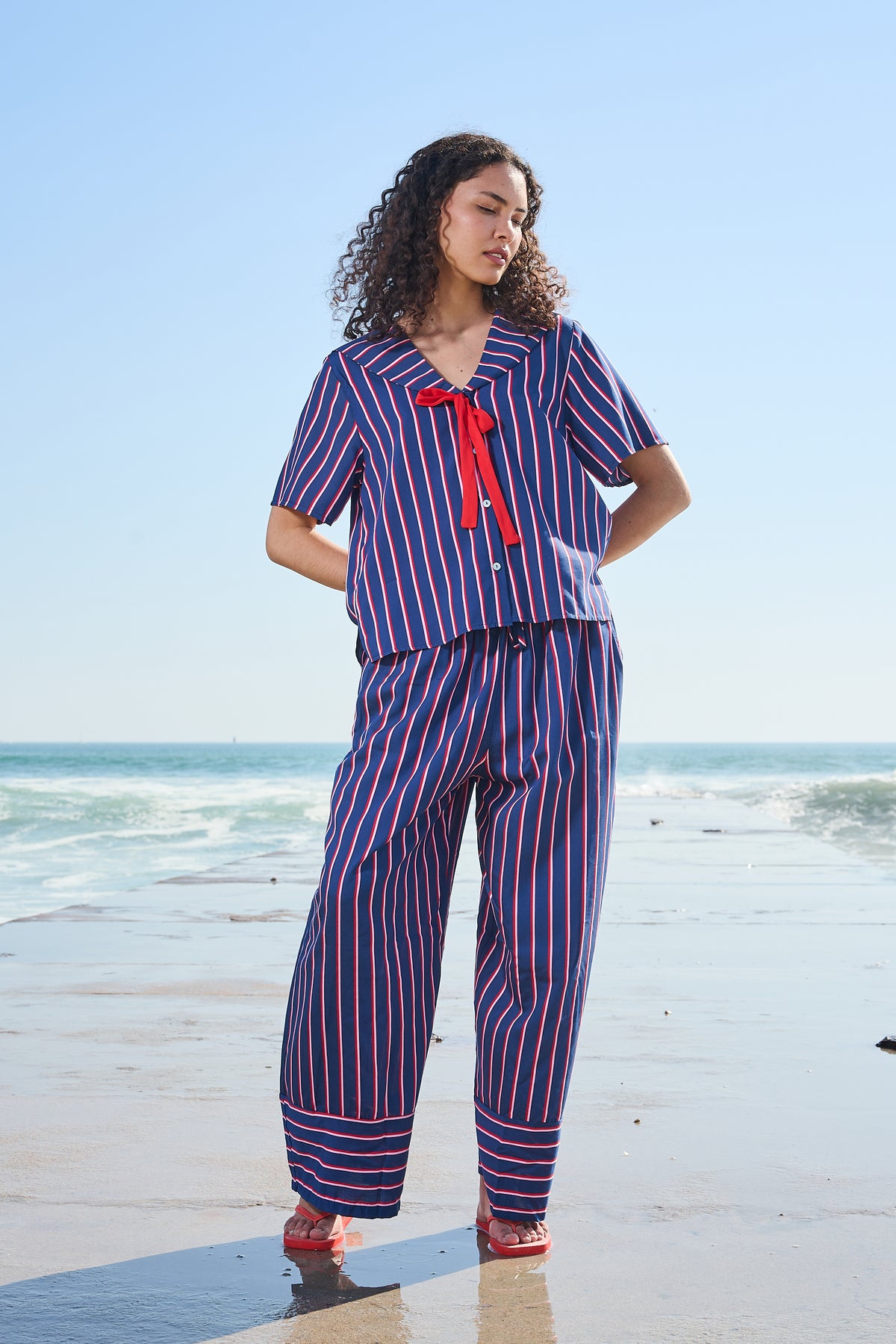 A woman stands on a beach wearing a matching two-piece set with vertical blue, red, and pink stripes, featuring a red bow tie detail on the short-sleeve top.