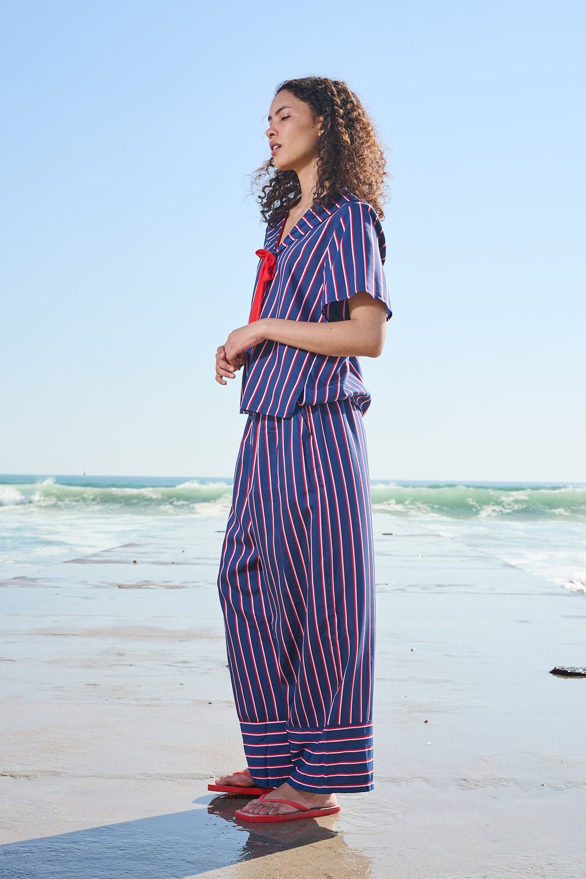 A woman stands on a beach wearing a matching two-piece set with vertical red and blue stripes, a red necktie, and red flip-flops.