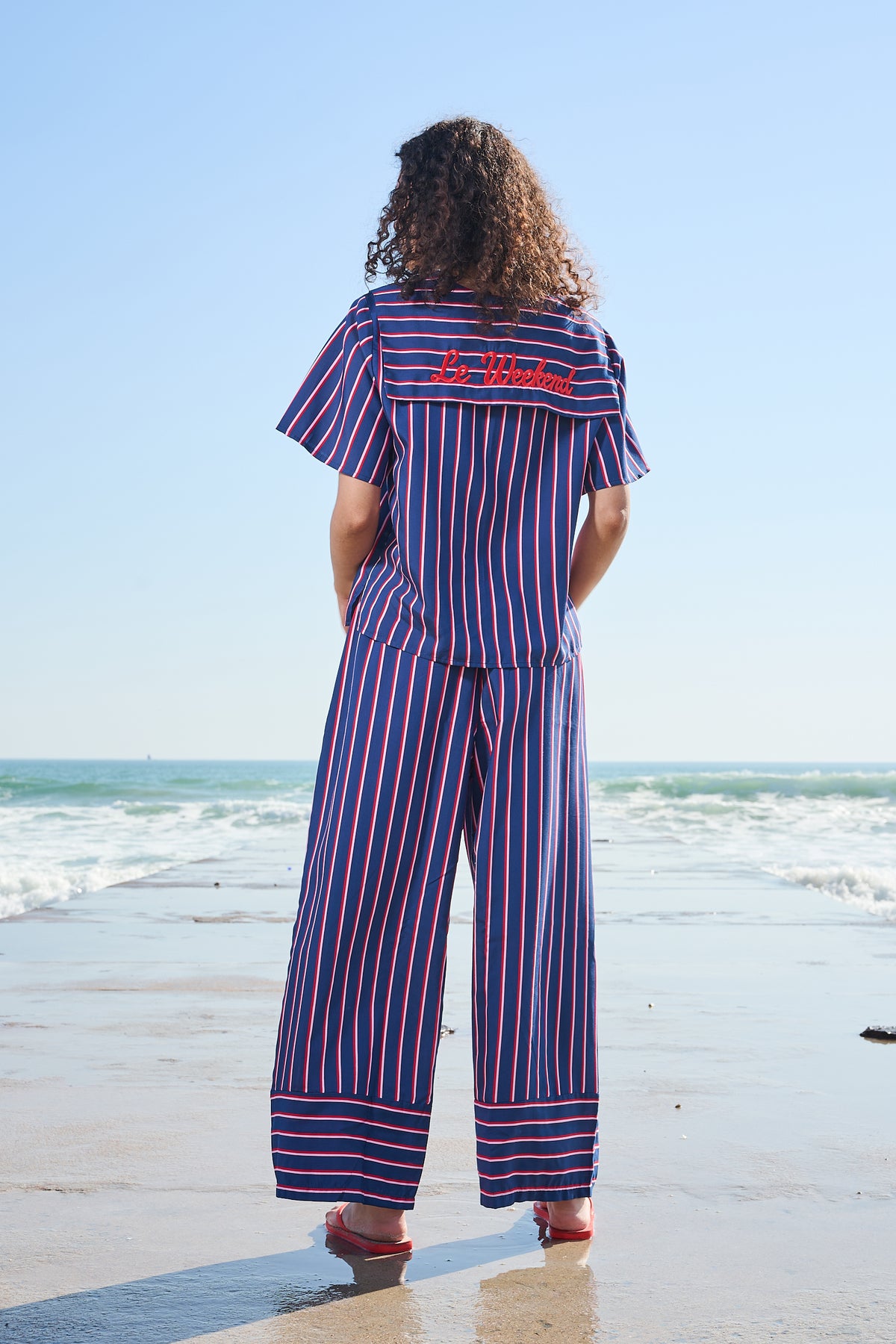 A person standing on a beach, viewed from behind, wearing a matching two-piece striped in navy blue, red, and white. The jumpsuit features a short-sleeve top and wide-leg pants, with 'Le Weekend' embroidered in red on the back.
