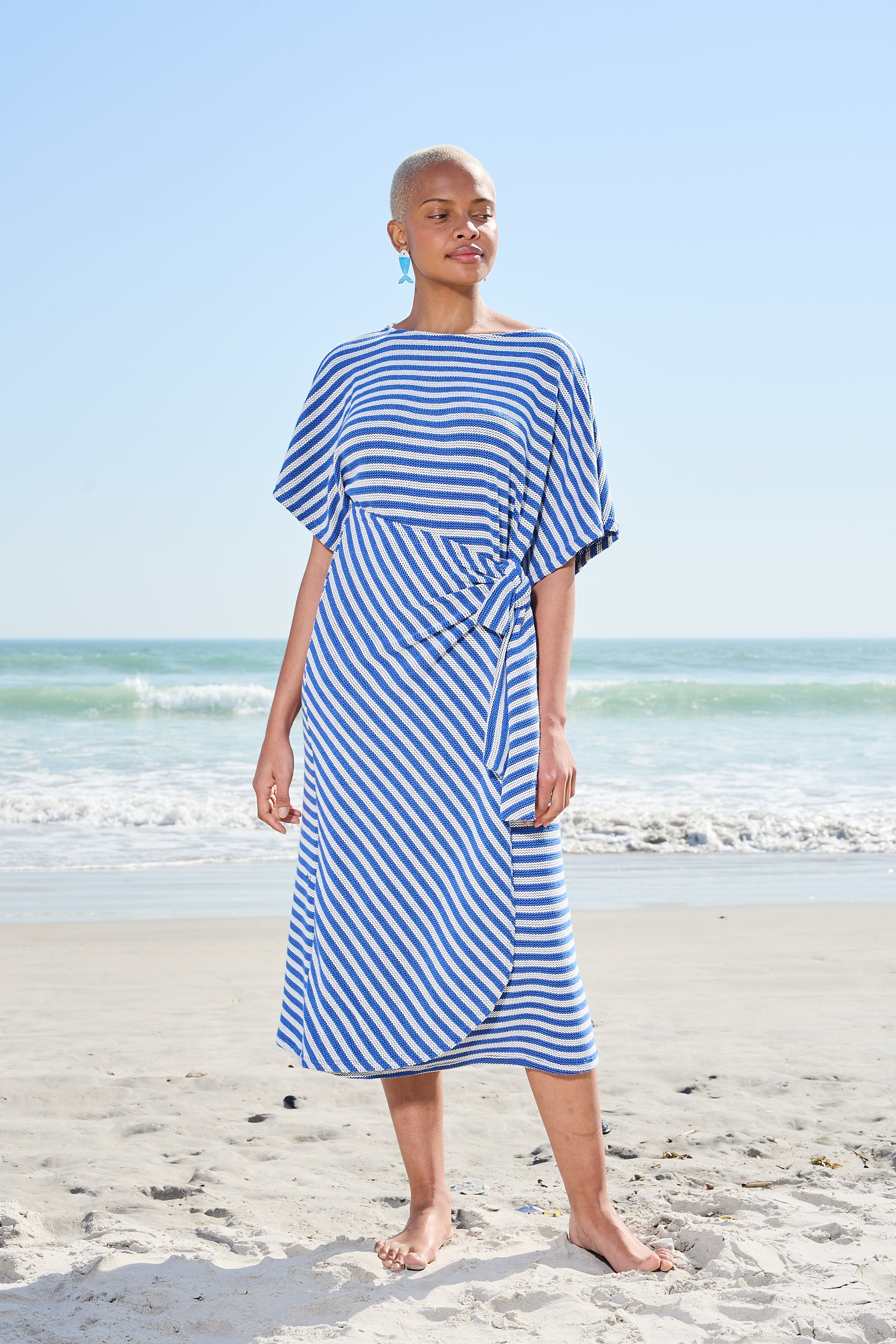 A woman wearing a blue and white striped midi dress stands on a sandy beach with the ocean and blue sky in the background.