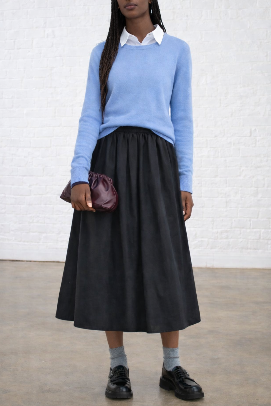 Woman wearing a light blue sweater, black skirt, and holding a purple bag against a white brick wall.