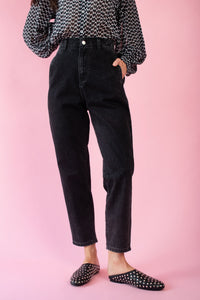 Model is wearing black denim mom jeans with a black and white blouse and black studded slip on shoes 