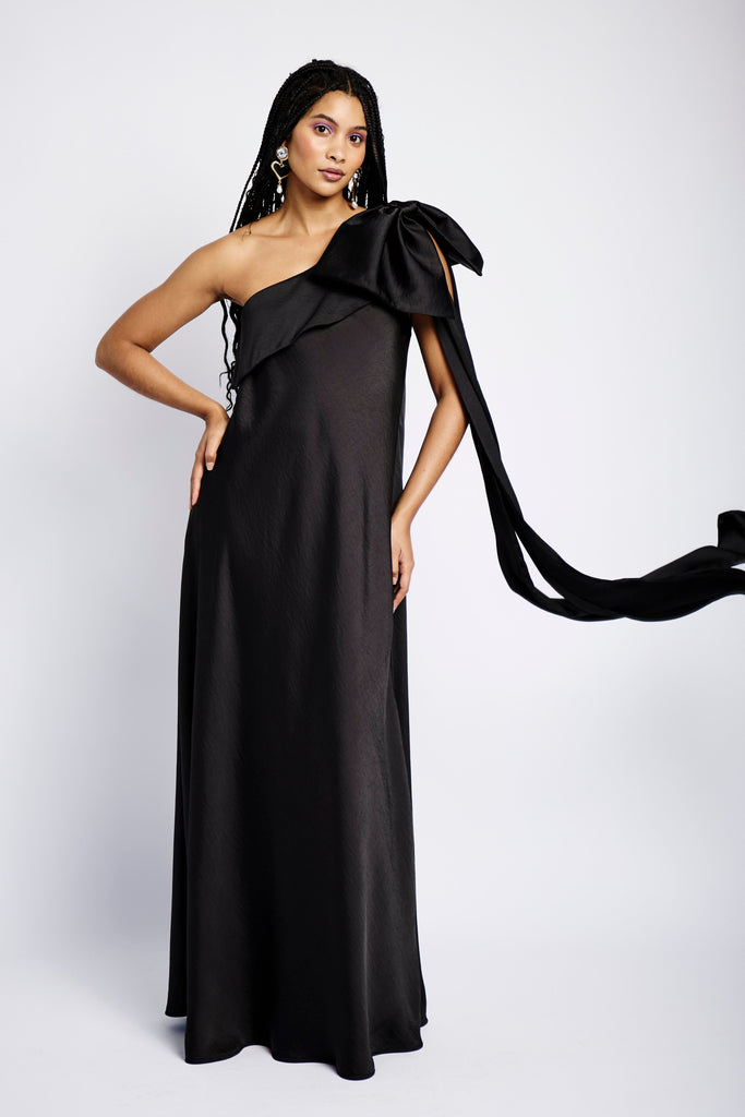 Model is wearing a black maxi satin dress with an asymmetrical neckline and a large bow on one shoulder