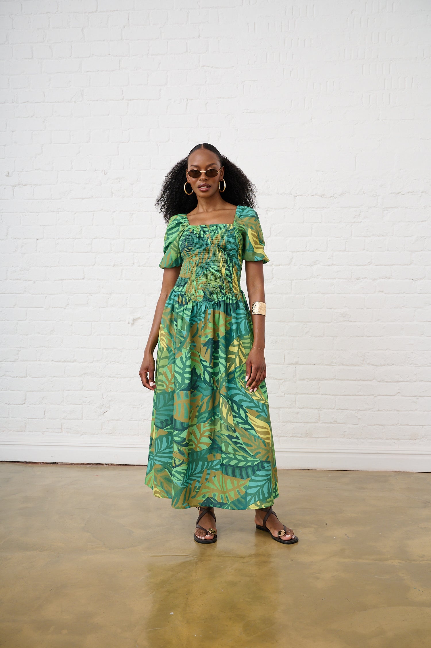 Woman wearing a green leaf-patterned dress standing against a white brick wall.