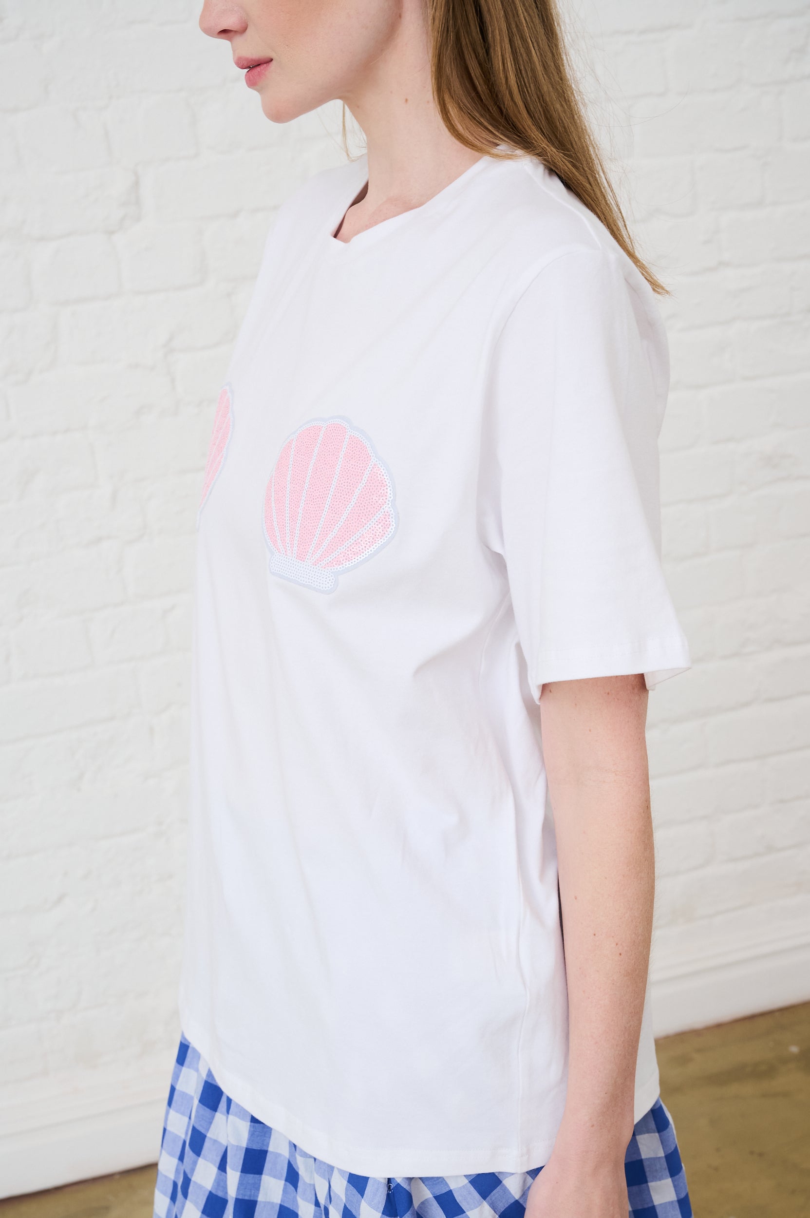 Person wearing a white t-shirt with two pink sequin shells on top against a white wall.