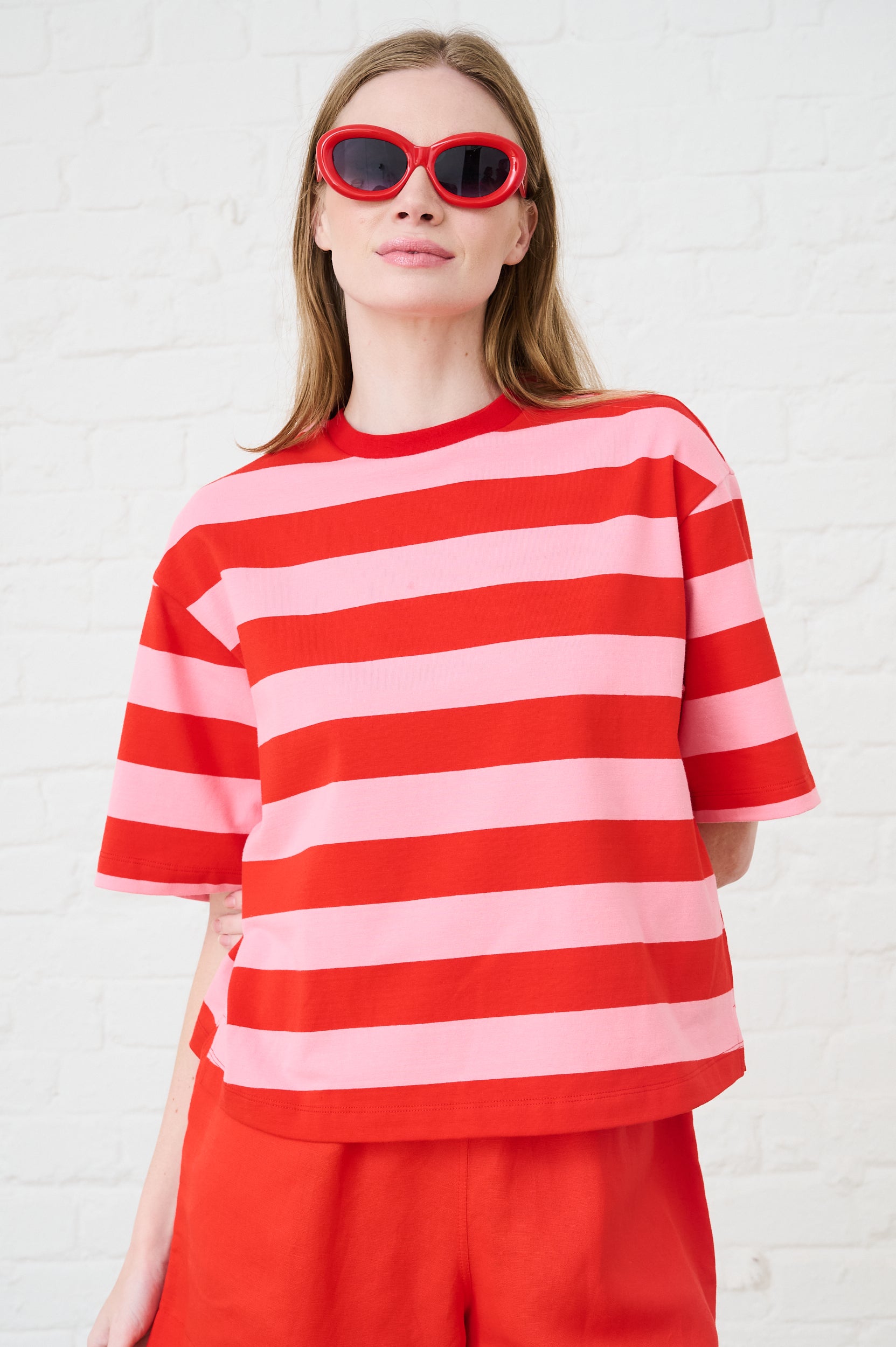 Model is wearing a pink and red stripe boxy tee with red sunglasses and red shorts