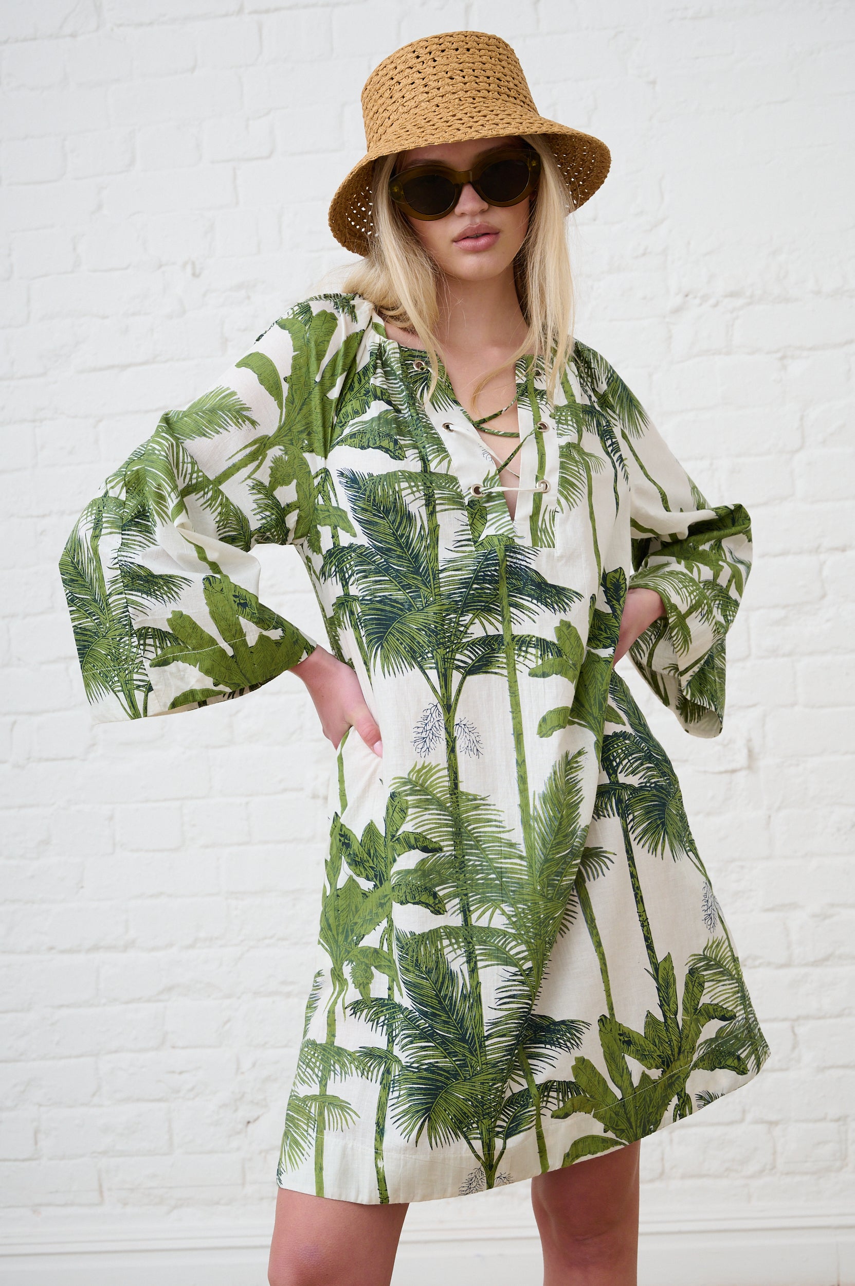 Model is wearing a 3/4 sleeve green and white palm tree print dress with a straw hat and sunglasses