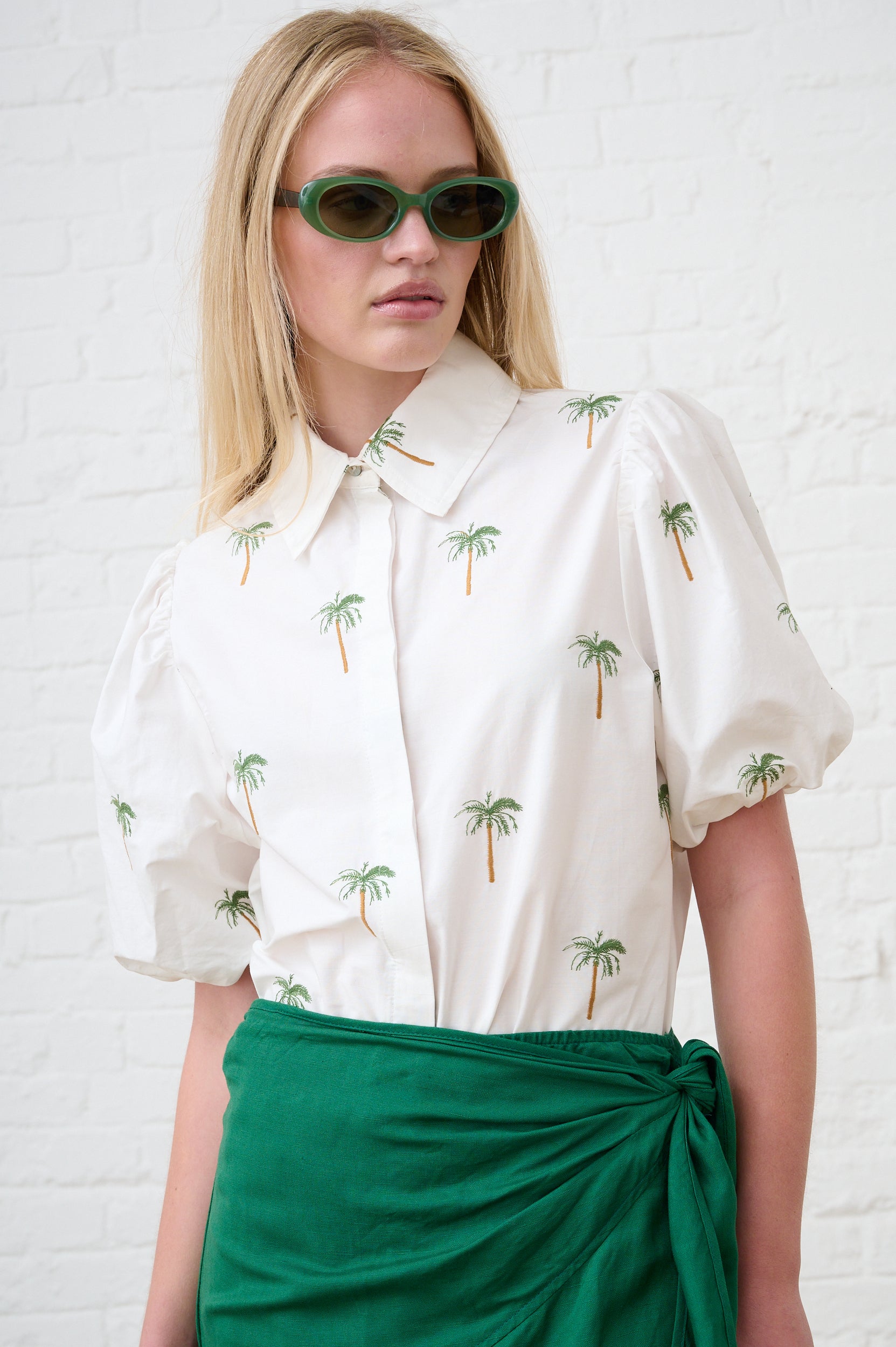 Model is wearing a white puffed sleeve shirt with embroidered palm trees on and a green wrap skirt 