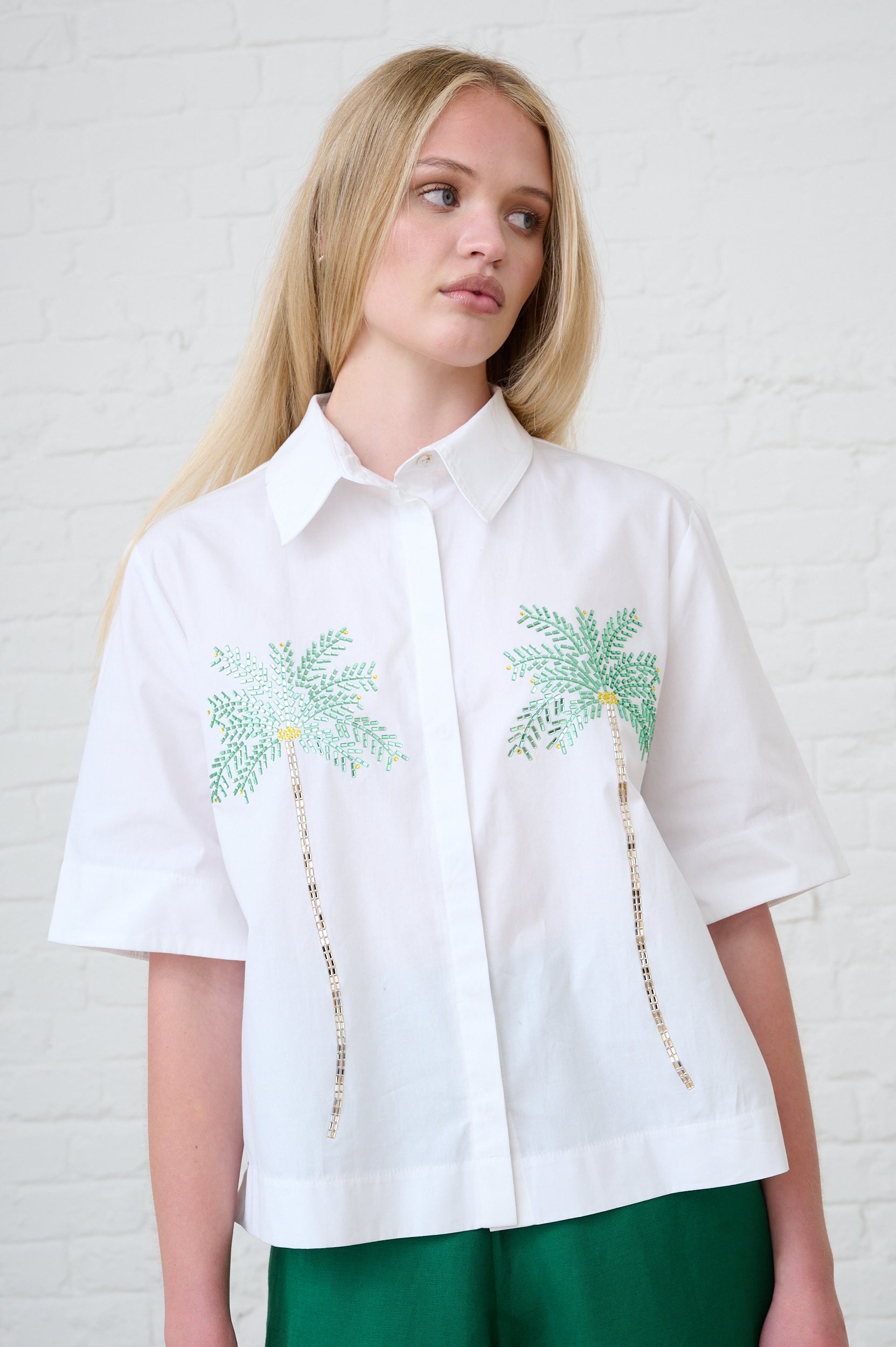 Woman wearing a white shirt with palm tree designs against a white brick wall.