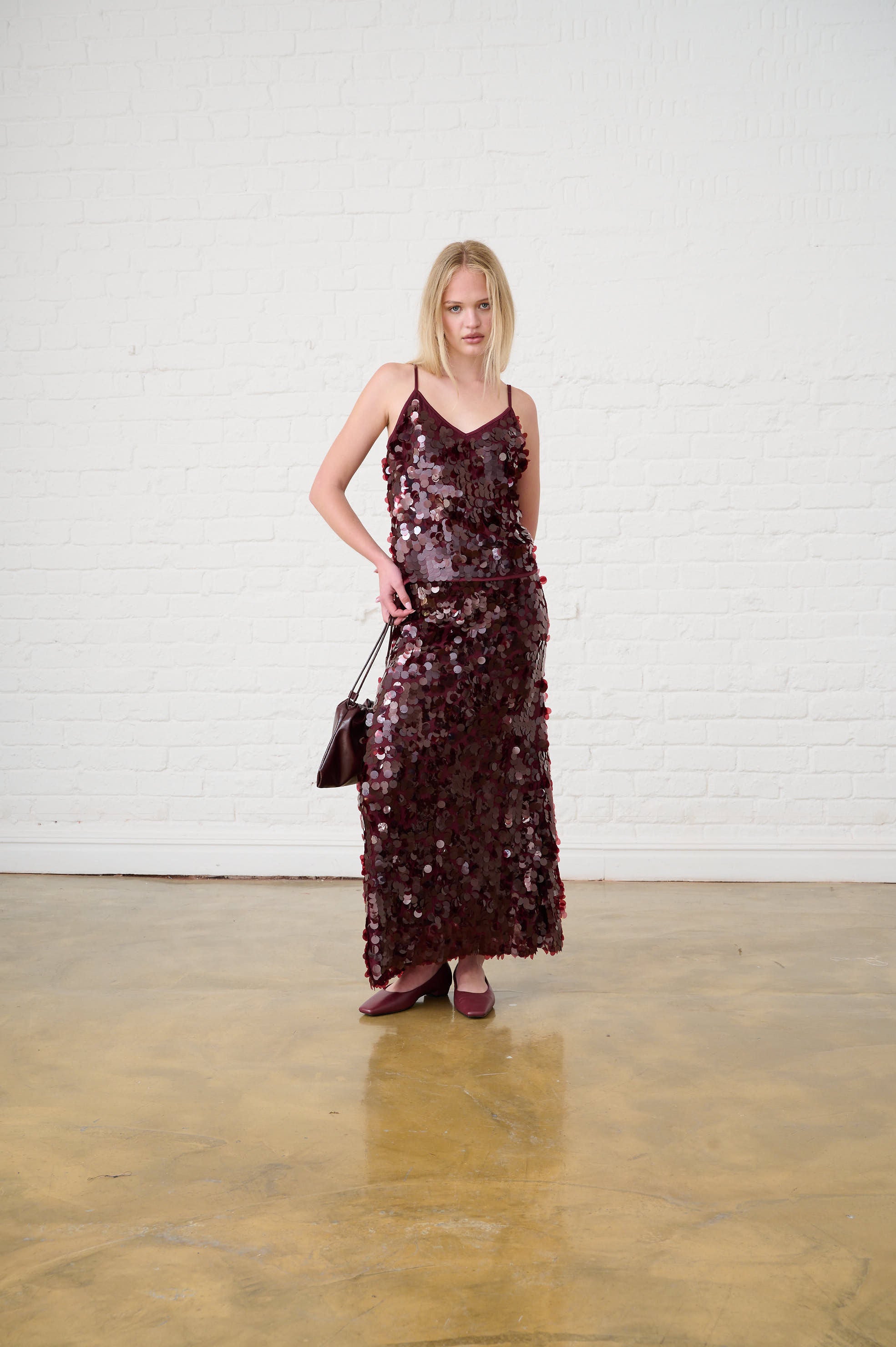 Woman wearing a sequin dress holding a handbag against a white wall.