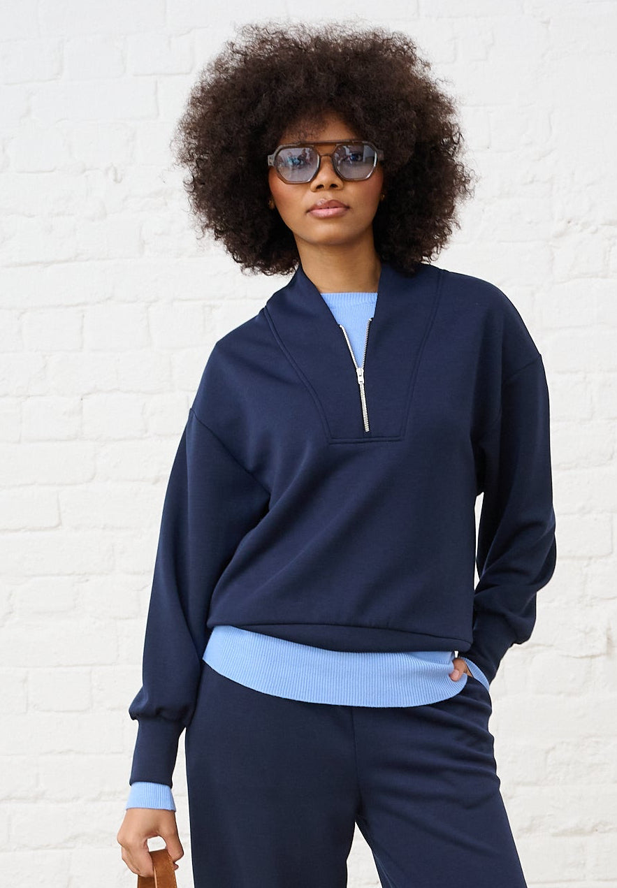 Model is wearing a navy quarter zip layered with a blue jumper and matching wide legs pants with leopard print sneakers  and brown accessories 