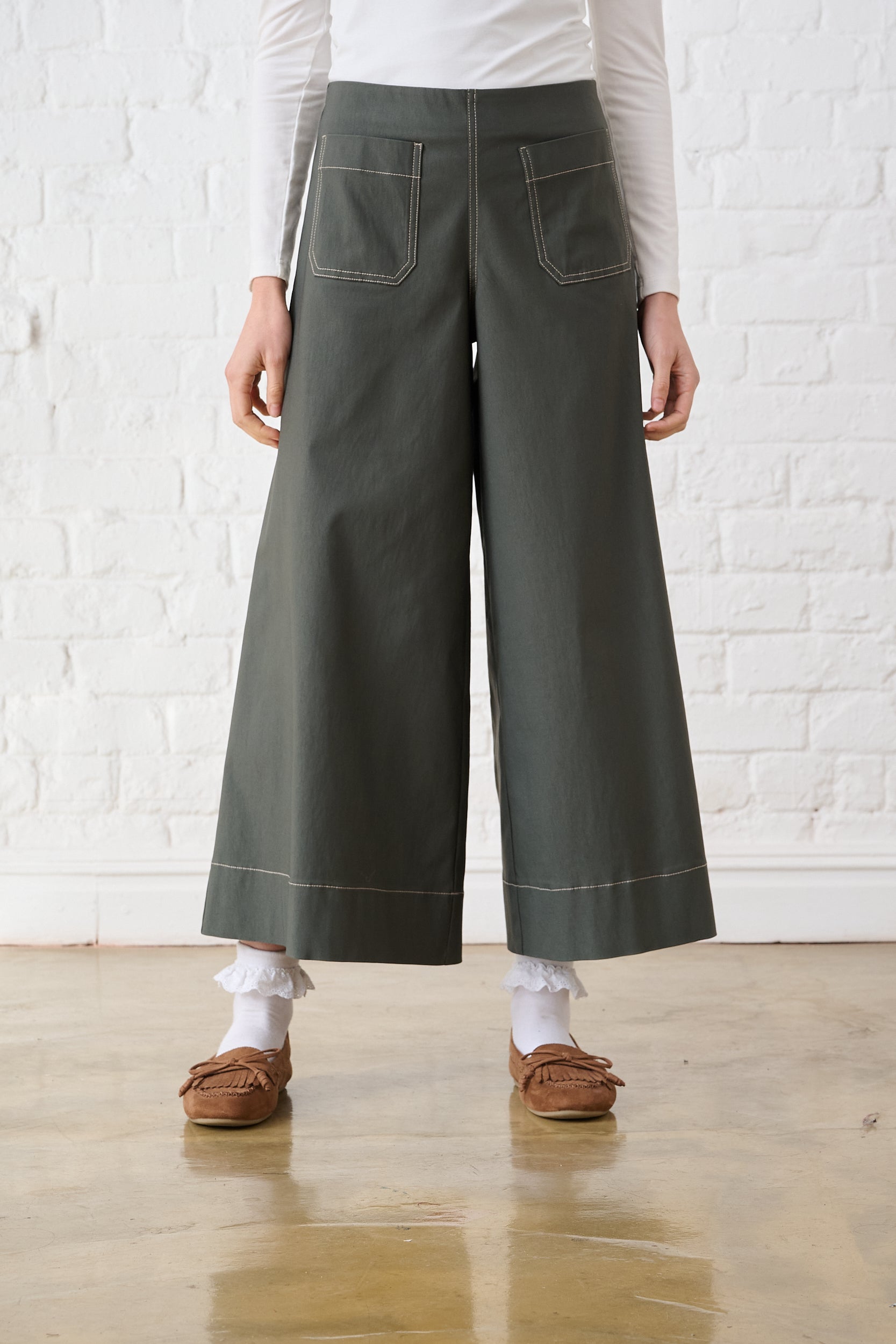 Model is wearing wide leg olive bengaline pants with contrast stitching and brown shoes 