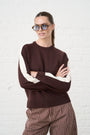 Woman wearing a brown sweater with white stripes and striped pants against a white brick wall.