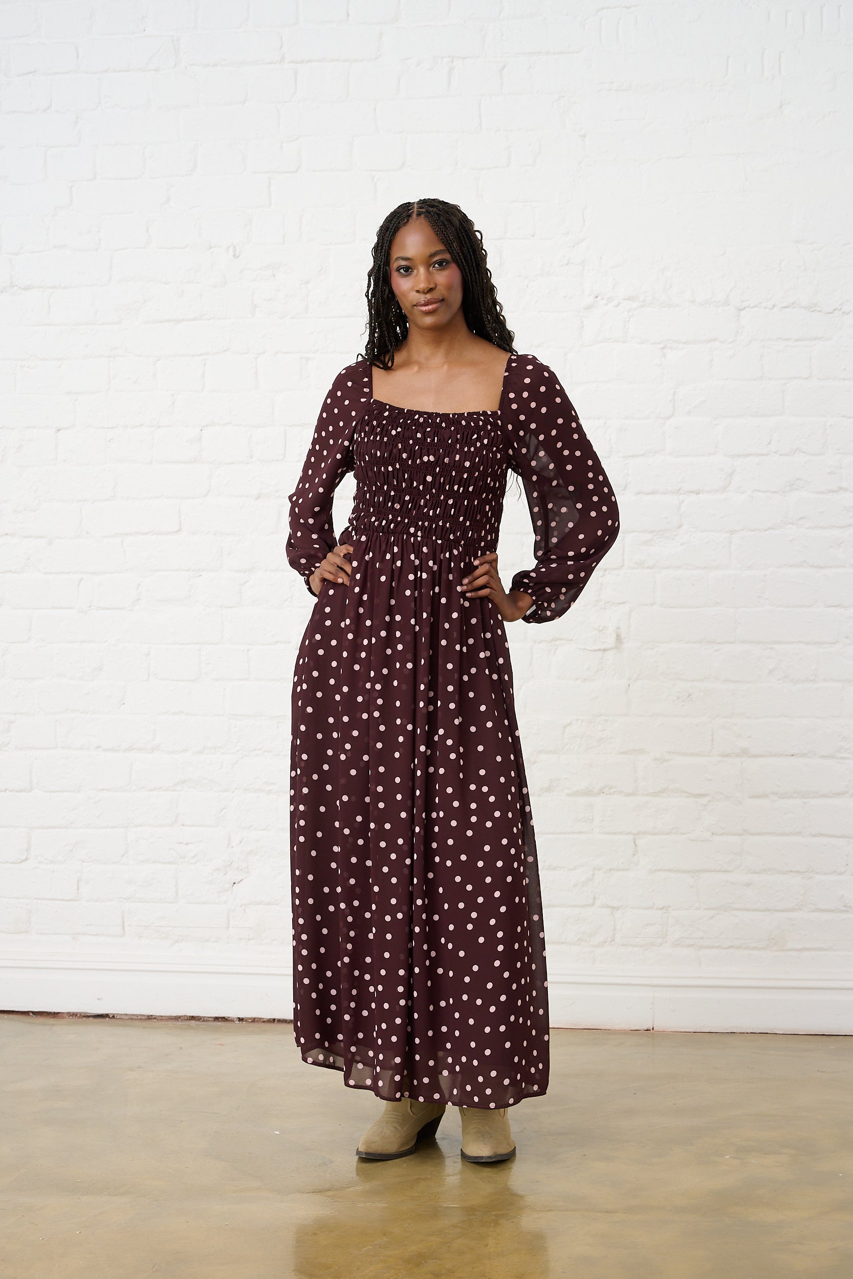 Model is wearing a burgundy maxi dress with pink polka dot print, shirred bodice, square neckline, long sheer sleeves and elasticated cuffs, styled with ankle boots.