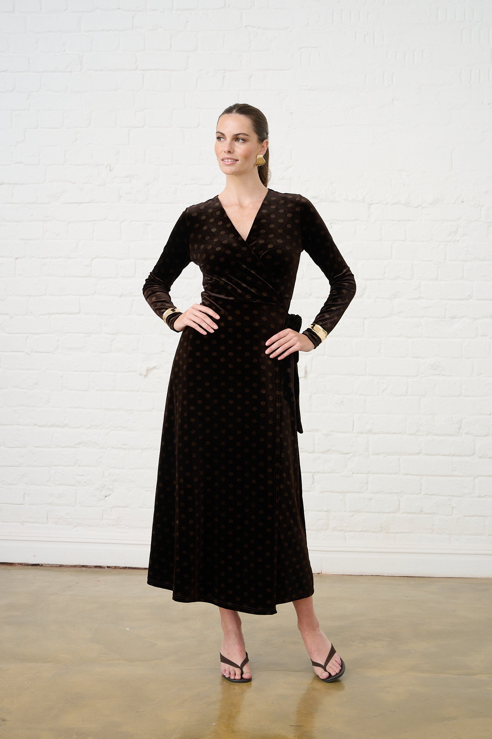 Model is wearing a brown velvet wrap dress in a spot print with gold accessories and black heels