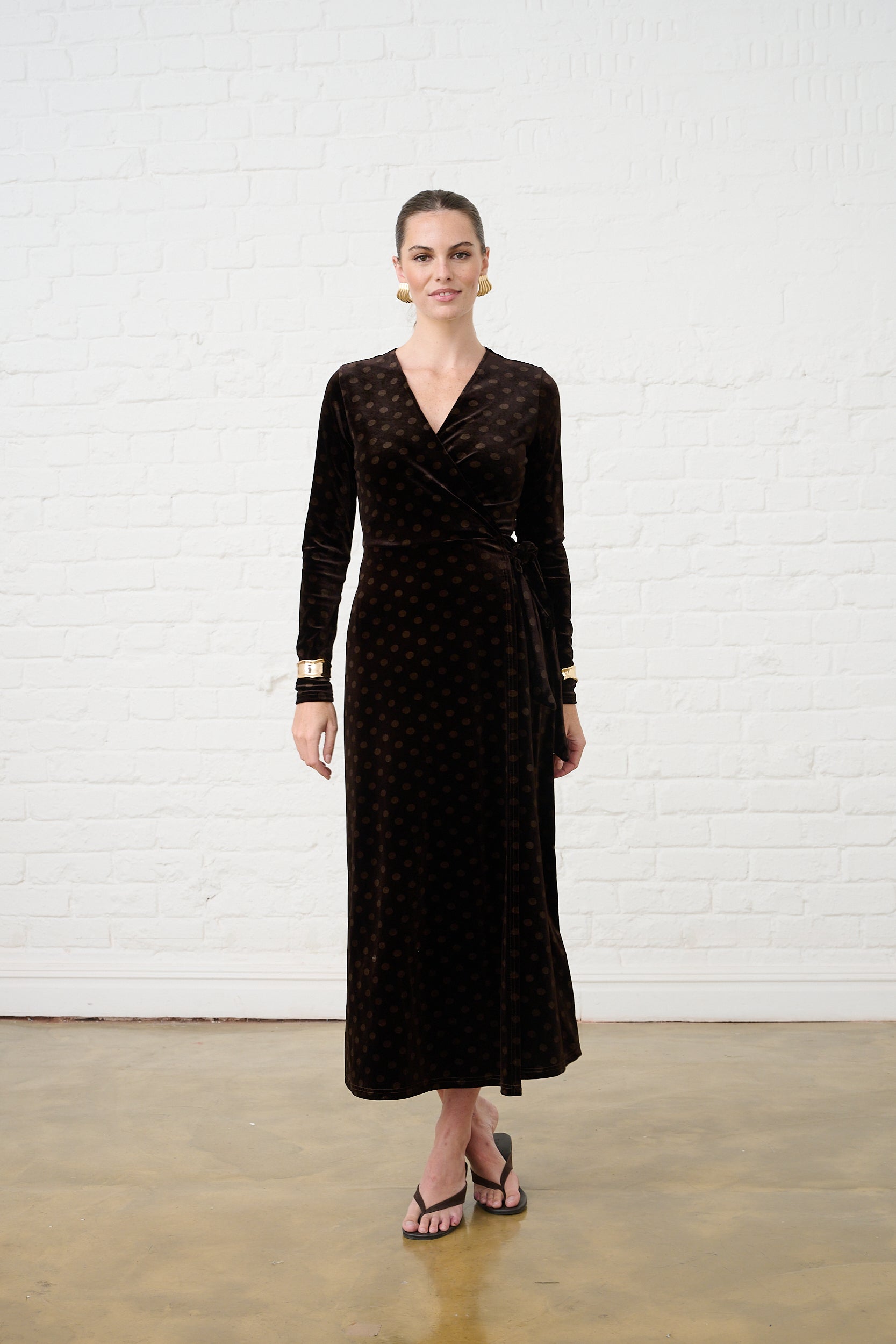 Model is wearing a chocolate brown tonal spot print velvet wrap dress with black heels and gold accessories 