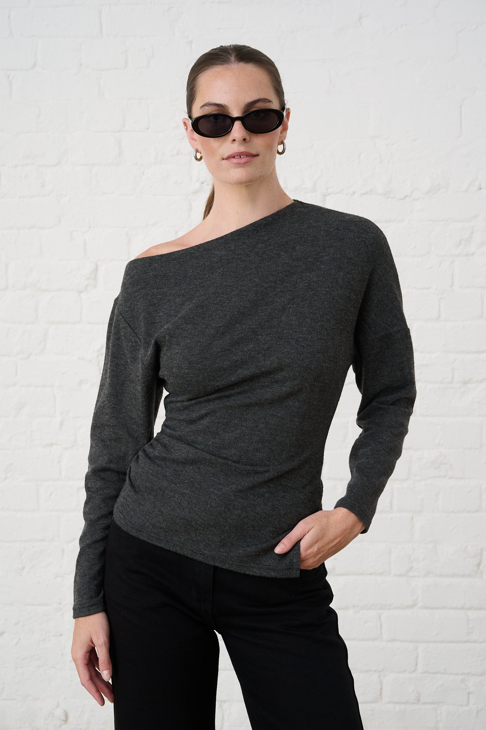 Model is wearing a grey asymmetrical long sleeve knit top with black denim jeans and black sunglasses 