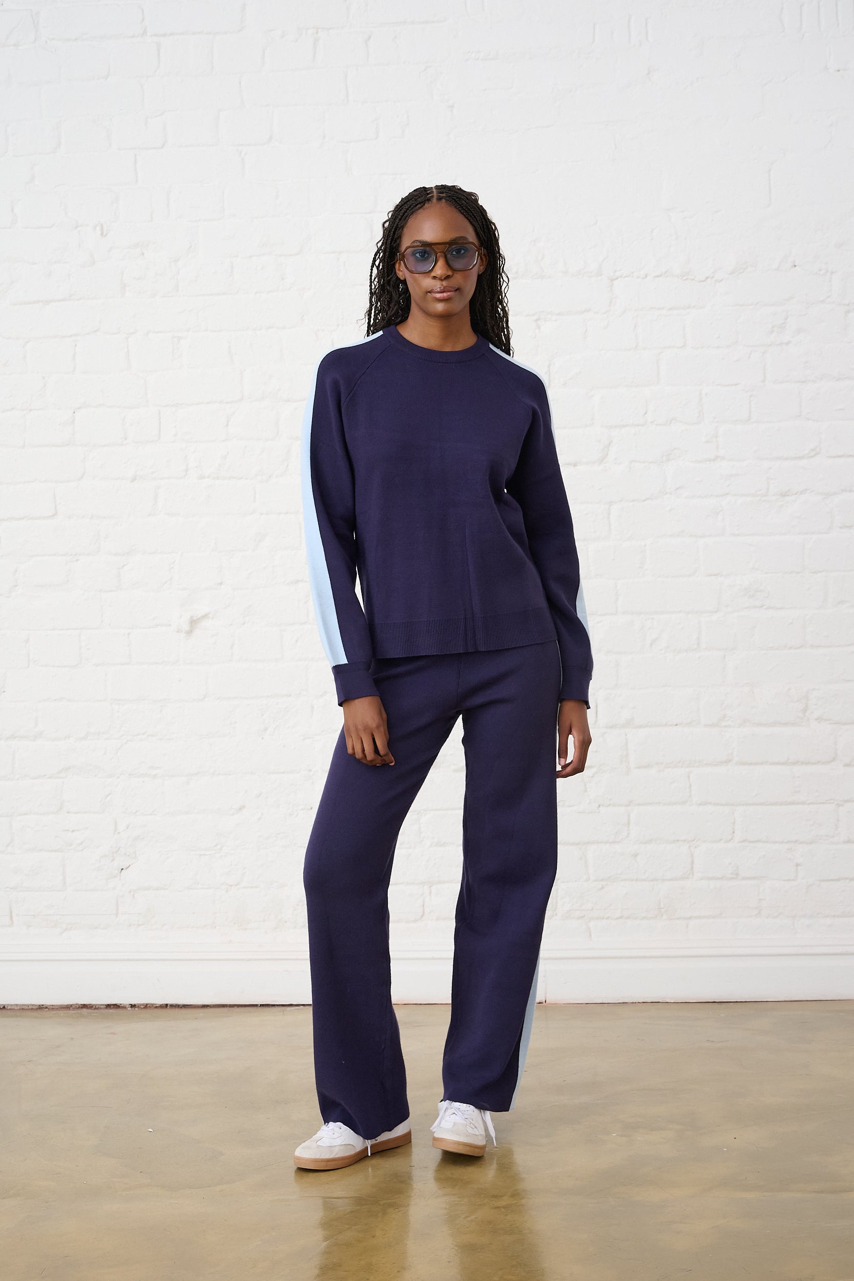 Model is wearing a blue knit set with light blue stripe detailing along the sides, with white sneakers and sunglasses