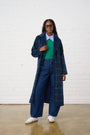 Model is wearing a long coat with green and navy check with a green vest and dark wash denim