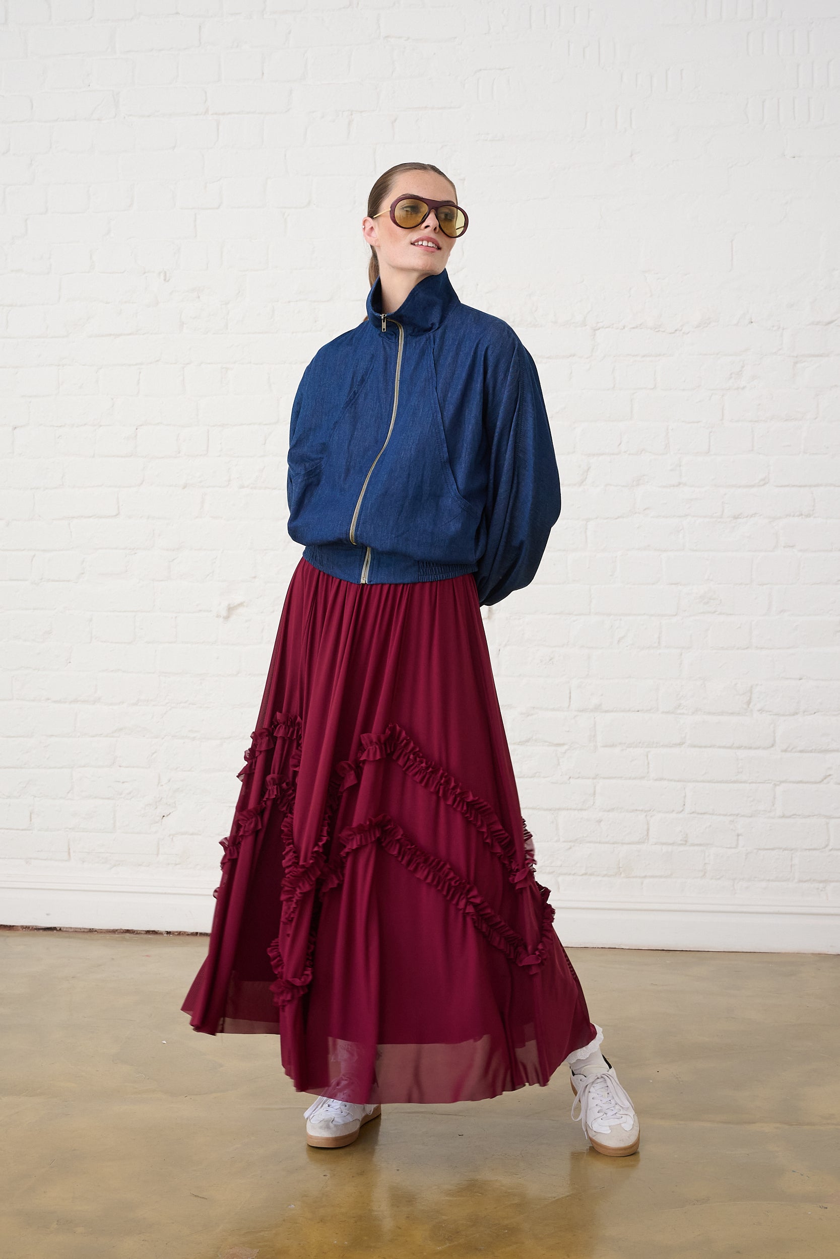 Model is wearing a denim bomber jacket with a burgundy mesh skirt and white sneakers 