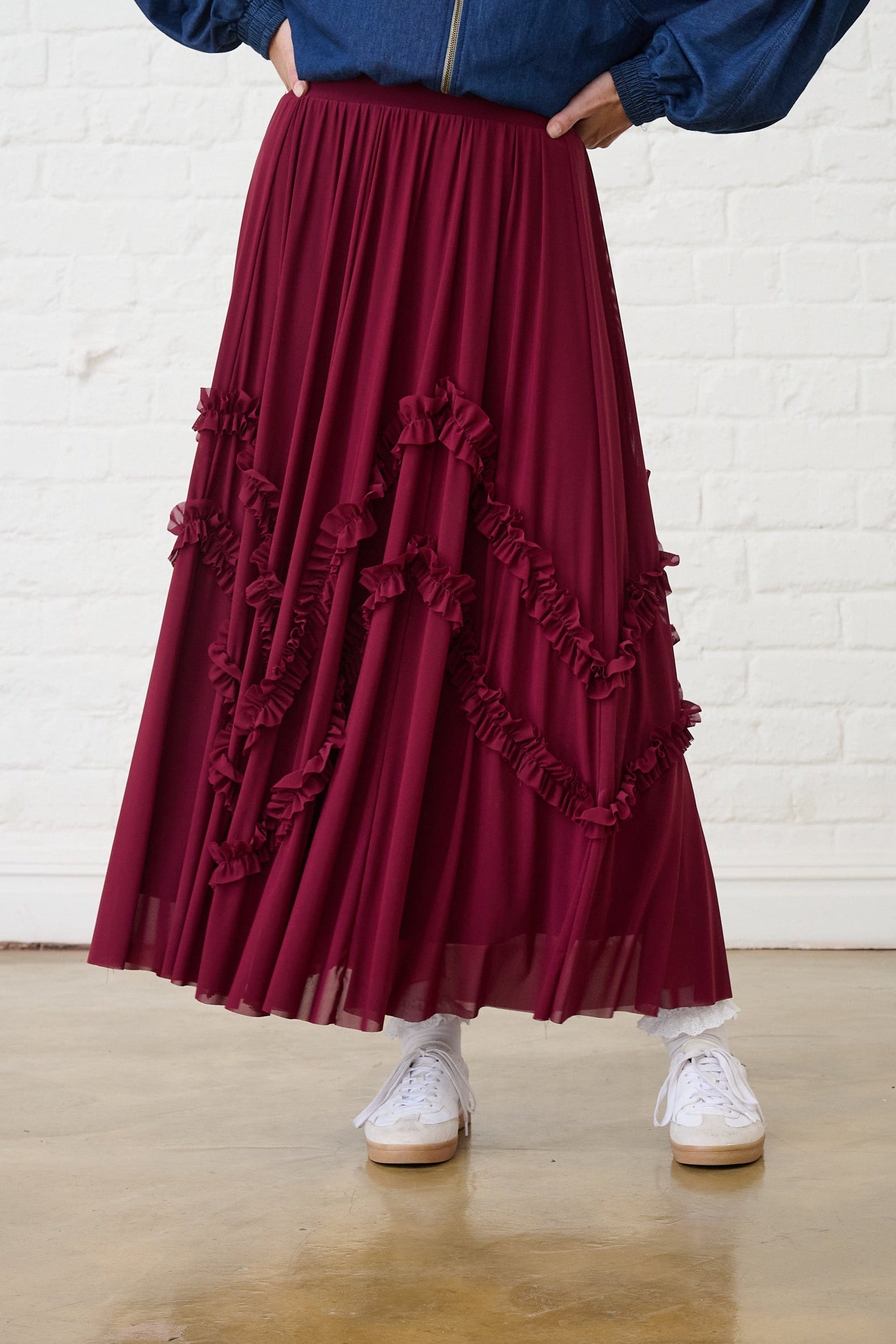 Model is wearing a burgundy ruffle skirt with a denim bomber and white sneakers 