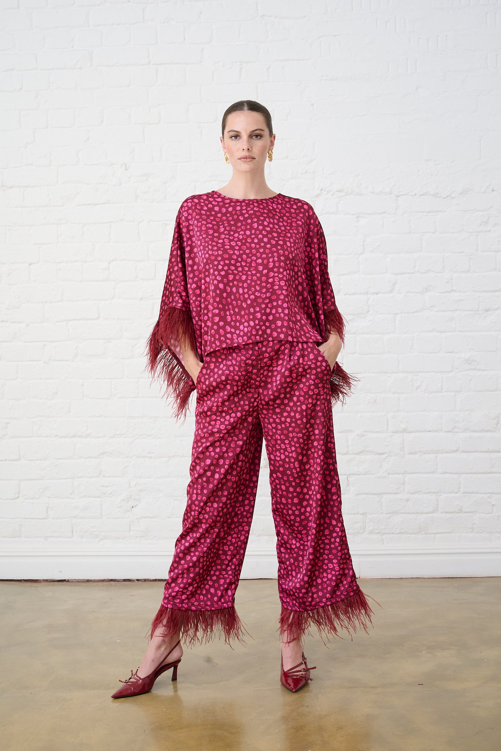 Model is wearing bright pink pants with a feather trim with a matching kimono top and burgundy shoes 