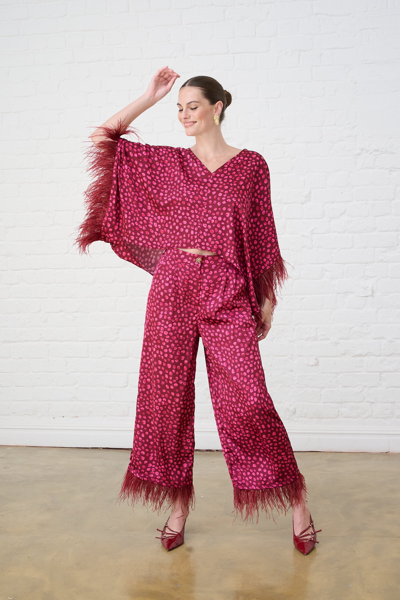 Model is wearing a bright pink kimono sleeve top with feather trim and matching pants with burgundy shoes