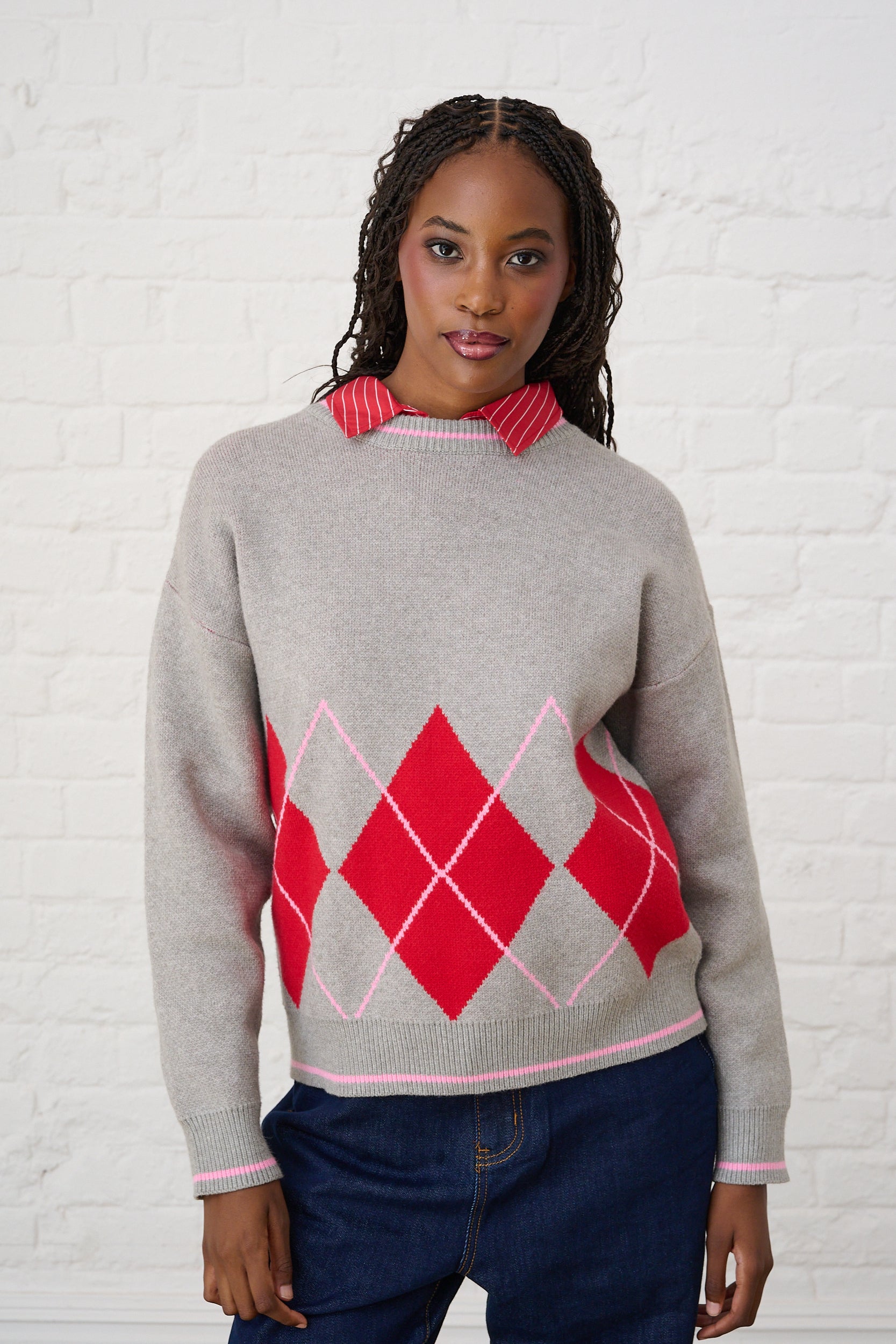 Model is wearing a grey knit sweater with red and pink argyle print, with dark wash denim jeans and a red shirt underneath 
