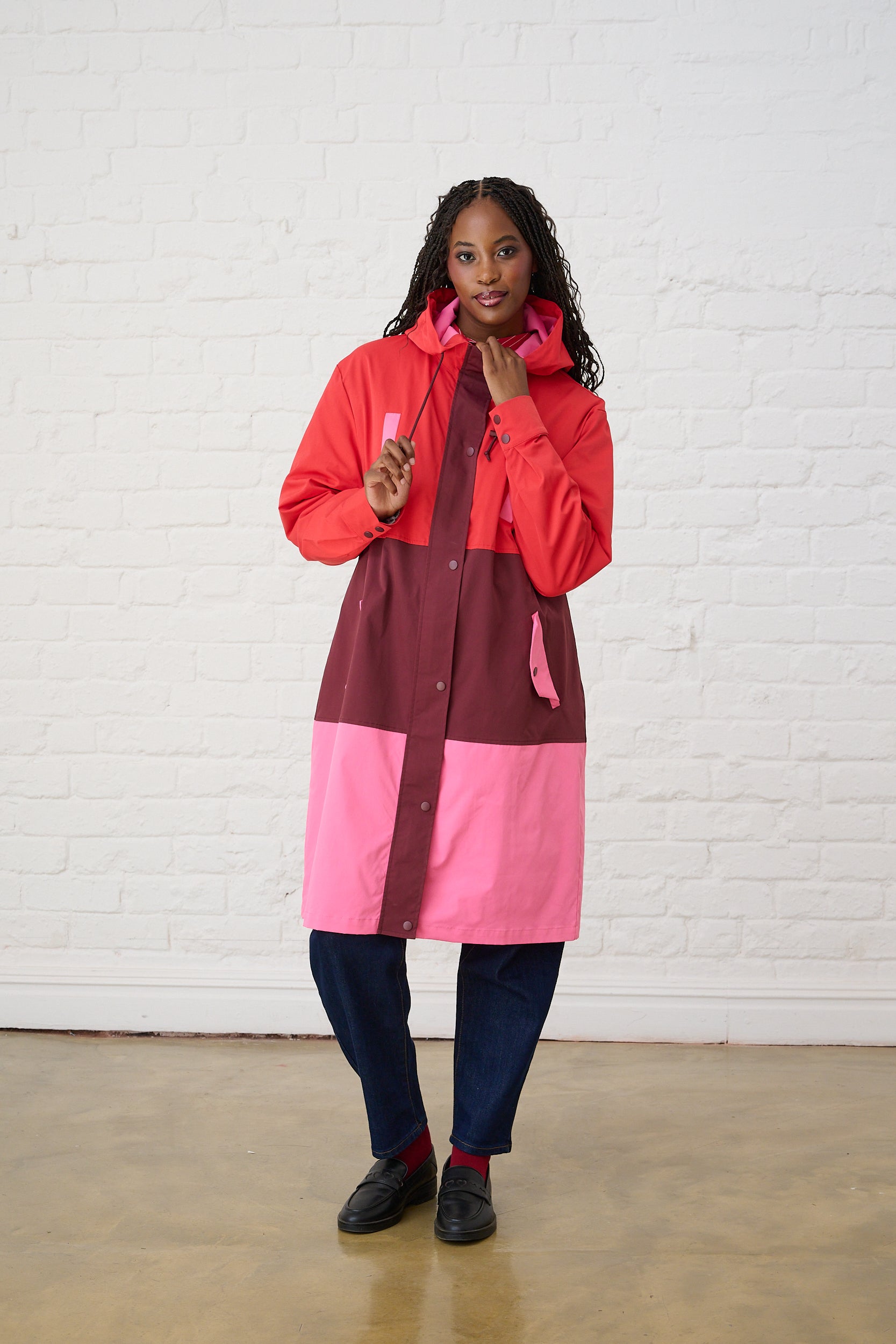 Model si wearing a colour block rain coat in pink and red with jeans and black loafers 