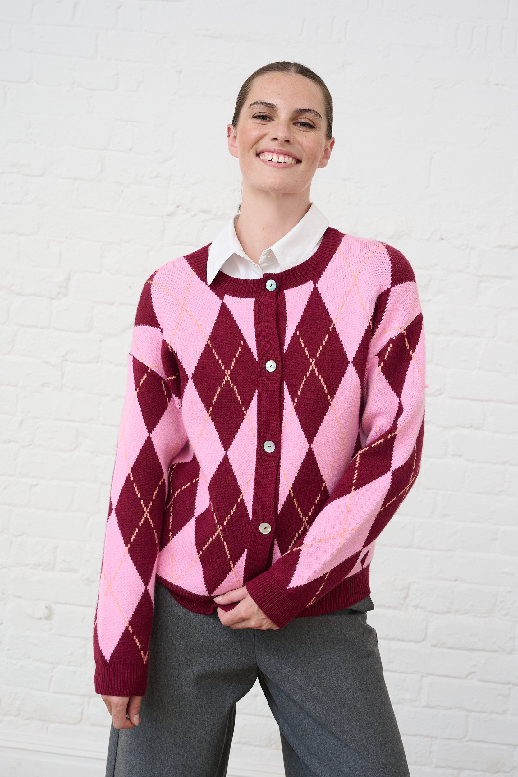 Model is wearing a burgundy and pink argyle button down cardigan with grey tailored pants and a white button down shirt layered underneath 