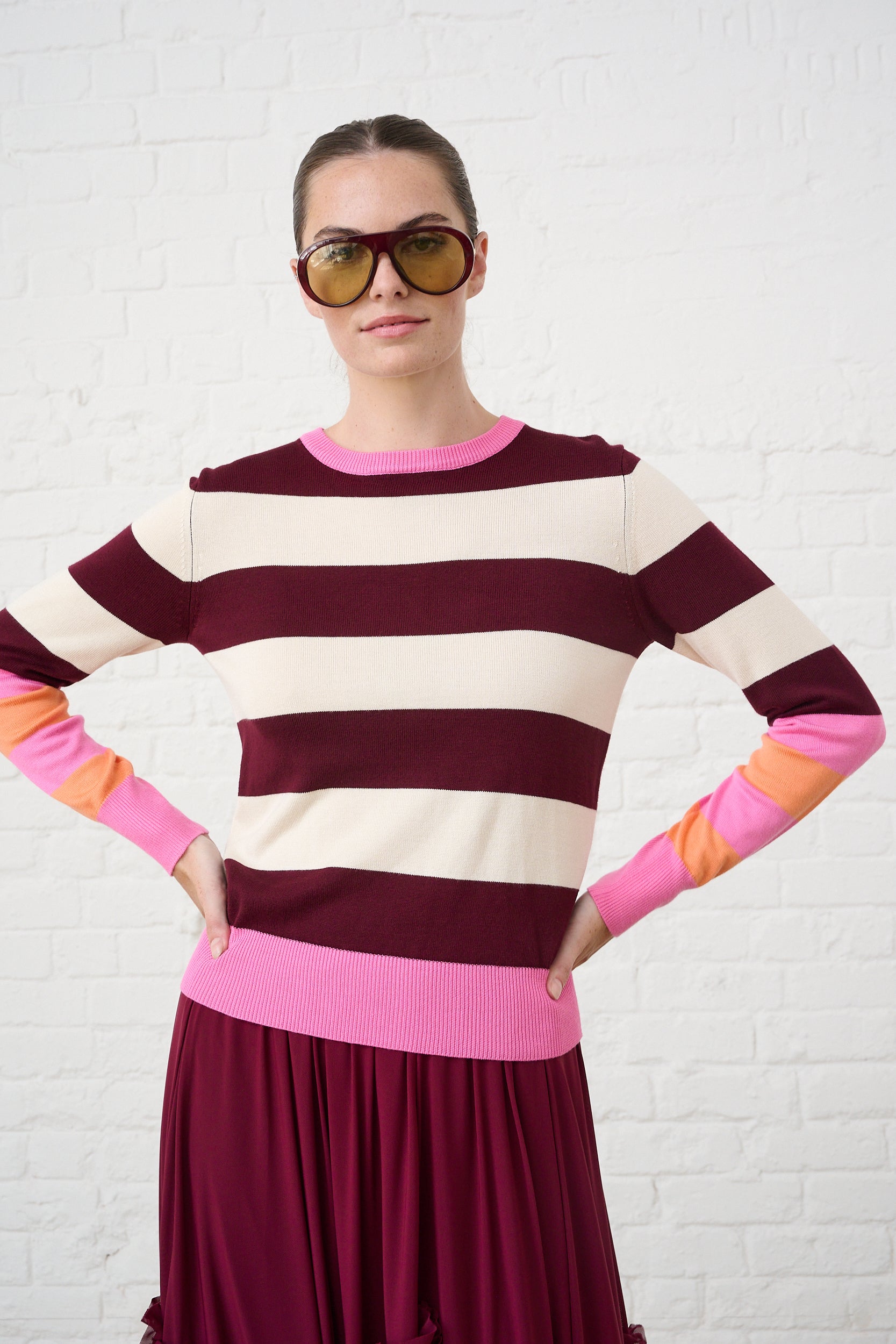 Model is wearing a burgundy, pink and orange bold striped jumper with burgundy mesh skirt 