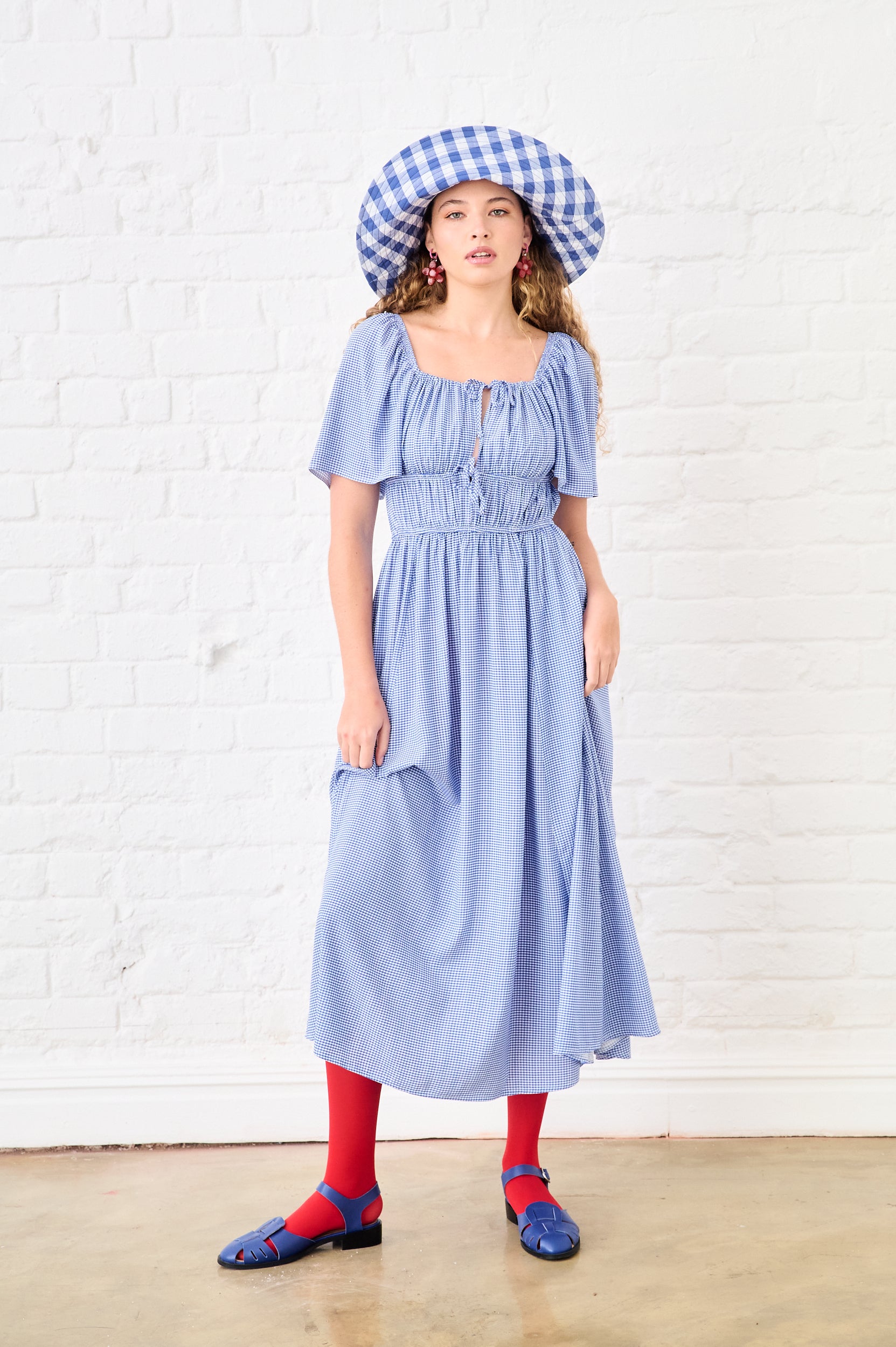 Woman wearing a blue dress and checkered hat against a white brick wall.