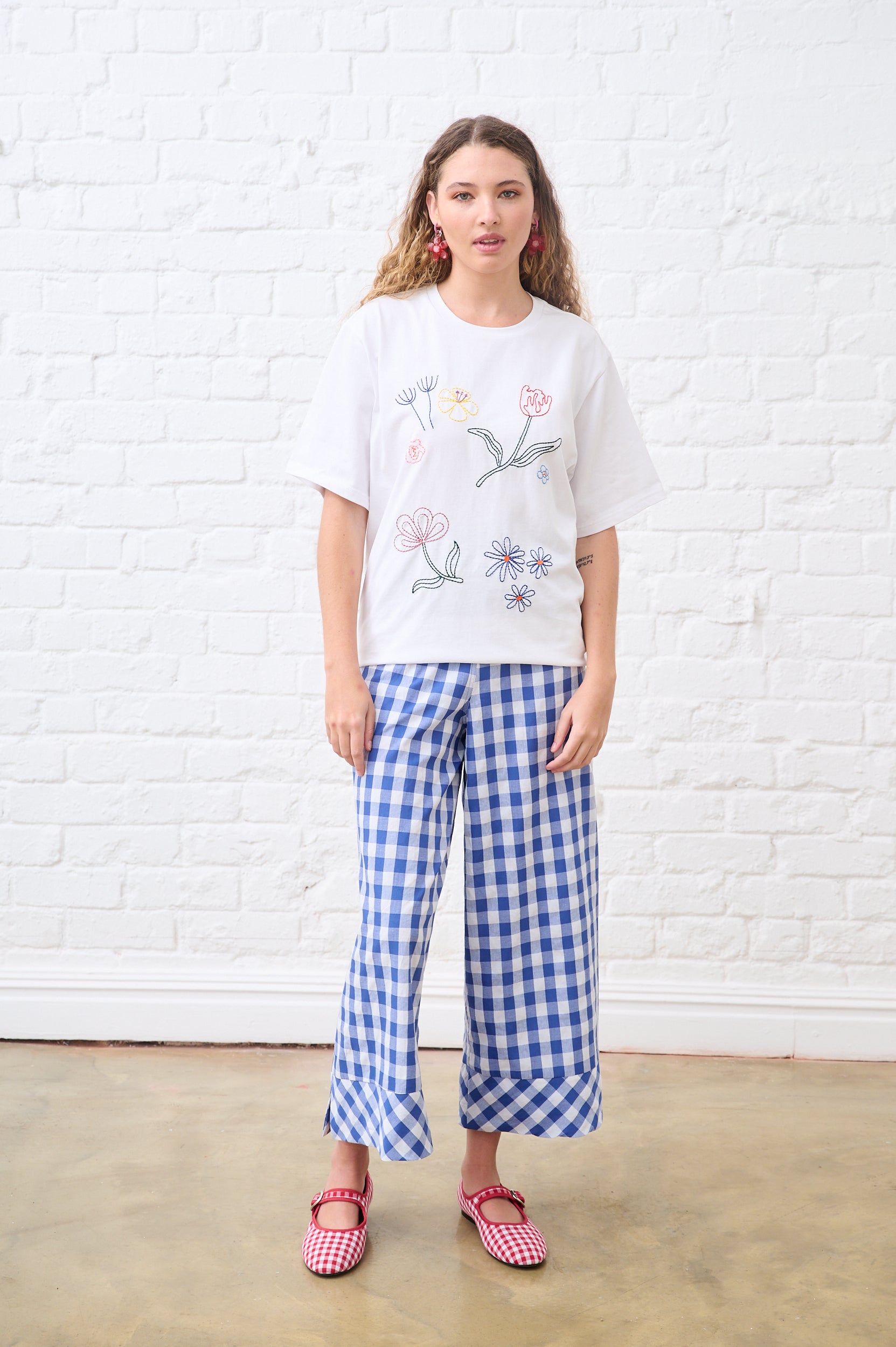 Woman wearing a white t-shirt with floral designs and blue checkered pants against a white brick wall.