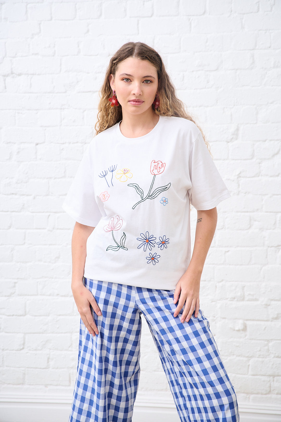 Woman wearing a white t-shirt with floral embroidery and blue checkered pants against a white brick wall.