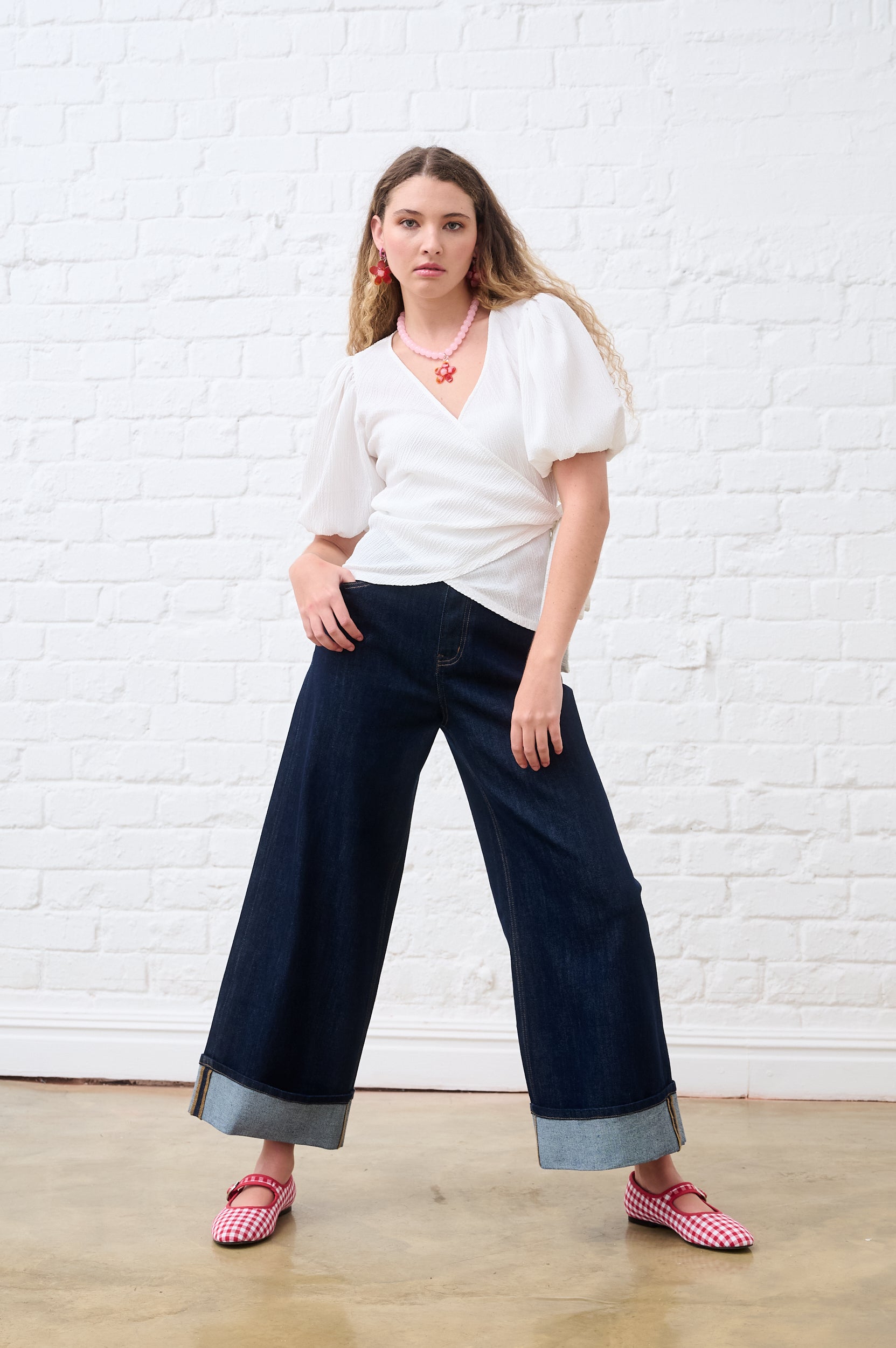 Woman wearing a white top and dark blue pants against a white brick wall.