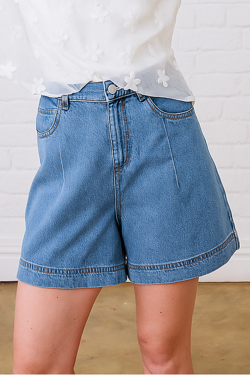 Woman wearing a white blouse with floral patterns and blue shorts against a white brick wall.